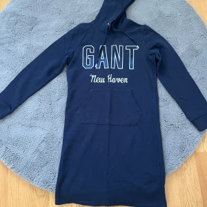 Marinblå hoodieklänning från GANT - Marinblå hoodieklänning från GANT med tryck 'GANT New Haven' på bröstet. Klänningen har lång ärm, huva med snörning och en stor magficka framtill. Skön och avslappnad passform, perfekt för chill dagar. Tillverkad i mjuk bomull.