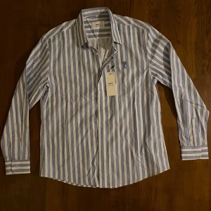 Randig skjorta  - Snygg skjorta  i vitt och ljusblått med vertikala ränder. Klassisk krage, långa ärmar och knäppning framtill. Broderad A-logga på bröstet. Tillverkad i mjuk bomull för en clean och fräsch look.