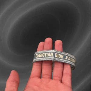 Grått armband Christian Dior J'Adior - Snyggt grått armband från Christian Dior med texten 'J'Adior' broderad i vitt. Armbandet har en stilren och modern design med diskret mönster längs kanterna. Perfekt för dig som vill ha en lyxig accessoar med ikonisk logga.