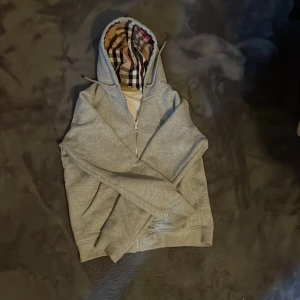 Grå Burberry hoodie med dragkedja - Säljer en grå hoodie från Burberry i storlek S Klassisk Burberry-rutig huva, broderad logga på bröstet och hel dragkedja framtill. Tillverkad i mjuk bomull med fickor på sidorna. Perfekt för chill dagar eller när du vill ha en stilren look.