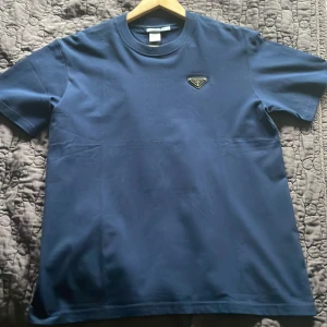 Mörkblå t-shirt från Prada - Snygg mörkblå t-shirt från Prada med klassisk passform och rund hals. T-shirten har ett diskret Prada-logomärke på bröstet och är tillverkad i mjuk bomull. Perfekt för dig som gillar stilrena och exklusiva plagg. Äkta fick den i julas men passar fortfarande inte mig e stor på mig o jag e 163. Passar nog 170-180. Den e S men passar även M