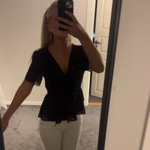Svart blus med v-ringning och volang - Säljer en svart blus med v-ringning och korta ärmar. Blusen har volang nedtill och är tillverkad i ett lätt, transparent tyg med diskreta prickar.  Tröjan är från newyorker och har klippt av armarna själv 