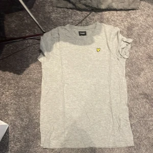 Grå t-shirt från Lyle & Scott - Snygg grå t-shirt från Lyle & Scott med klassisk rund hals och korta ärmar. På bröstet sitter den ikoniska gula örnen som broderad detalj. T-shirten är gjord i mjuk bomull och har en clean, enkel look som funkar till jeans eller shorts.