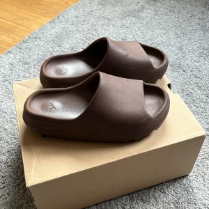 Adidas Yeezy Slide Flax strl 40 men passar 37-38 - Säljer ett par Adidas Yeezy Slide i färgen Flax. Slidesen är bruna, har en chunky och ribbad sula samt öppen tå. Tillverkade i syntetmaterial och har en clean, minimalistisk look med Yeezy-logga på insidan. Perfekta för chill dagar eller streetwear-stil. Kommer med box, dom skönaste skorna jag någonsin haft på mig känns som att man går på moln 
