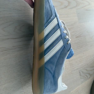 Adidas Samba i ljusblå mocka - Säljer ett par Adidas Gazelle sneakers i snygg ljusblå mocka med vita detaljer och klassiska tre ränder på sidan. Skorna har beige aktig sula, vit snörning och blå plös med Adidas-logga. Perfekta för dig som vill ha snygga varjedags skor.