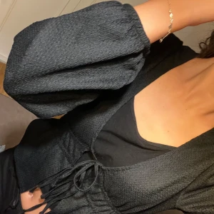 Svart kofta med snörning - Snygg svart kofta med långa puffärmar och markerad midja med snörning framtill. Koftan har en djup v-ringning och är gjord i ett strukturerat, mjukt material som ger en cool look. Perfekt för dig som gillar stilrena och trendiga plagg.