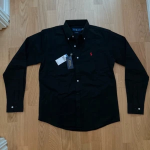 Svart skjorta från Ralph Lauren - Klassisk svart skjorta från Ralph Lauren med röd broderad logga på bröstet. Skjortan har lång ärm, vita knappar och är tillverkad i bomull. Snygg och stilren modell som passar perfekt till jeans eller chinos.