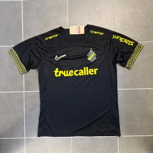 AIK svart matchtröja Nike 2024 Oanvänd - AIK:s officiella matchtröja från Nike i svart med gula detaljer och tryck. Kortärmad modell med klubbmärke på bröstet, sponsorer som Truecaller och SVEA, samt mönstrade muddar i gult. Tillverkad i lätt och ventilerande polyester, perfekt för fotboll och supporterhäng.