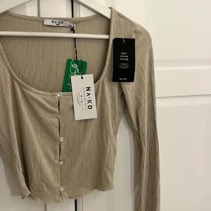 Beige ribbad topp från NA-KD - Trendig beige ribbad topp från NA-KD med croppad passform och långa ärmar. Toppen har snygg knäppning framtill med små knappar och en djup, rundad urringning. Perfekt att styla med jeans eller kjol för en cool vardagslook. Storleken är XXS men den passar S 