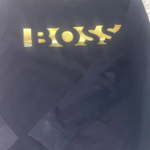 Svart BOSS tröja med guldfärgat tryck - Svart långärmad tröja från BOSS med ett stort guldfärgat BOSS-tryck på bröstet. Klassisk rund halsringning och ribbade muddar vid ärmslut och nederkant. Perfekt för dig som gillar stilrena och snygga loggoplagg.