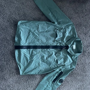 Grön overshirt från C.P. Company - Snygg grön overshirt från C.P. Company med svart dragkedja framtill och klassisk krage. Jackan har flera fickor, inklusive en ikonisk ficka med linsdetalj på ärmen. Tillverkad i bomull och perfekt för lager-på-lager-stil.
