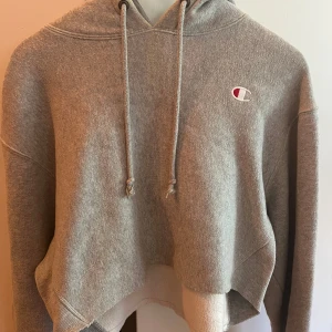 Grå croppad hoodie från Champion - Grå croppad hoodie från Champion med broderad logga på bröstet. Tröjan har huva med snörning och är tillverkad i mjuk bomullsblandning. Perfekt för en chill och sportig look. Reverse Weave-modell med avslappnad passform och långa ärmar.
