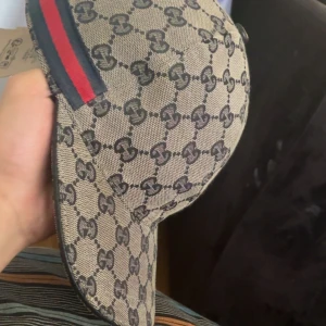Gucci beige keps med GG-mönster - Säljer en beige keps från Gucci med klassiskt GG-mönster i mörkgrått. Kepsen har en böjd skärm och en snygg detalj med röd och marinblå rand på sidan. Materialet är bomull och polyester, insidan är mörkblå. Perfekt för dig som gillar lyxiga accessoarer.