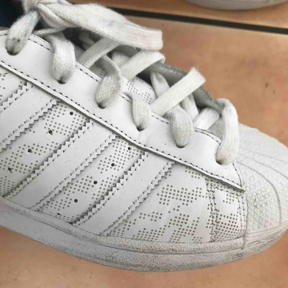 Originella Adidas Superstar som är vita och blåa med ett diskret tryck på. Innersulan är lite smutsig men går enkelt att byta ut. Blåa detaljer där bak som blir som reflexer med flash. Storlek 39 1/3 . Skor.