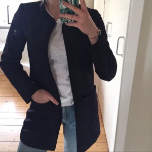  - Mörkblå öppen kappa med två fickor framtill. Storlek 36 och går till halva låret på mig som är ca 170. En söm är dragen på ett konstigt sätt (bild tre) men inget jag har lagt märke till tidigare. Kappans färg kommer fram på bild tre. 