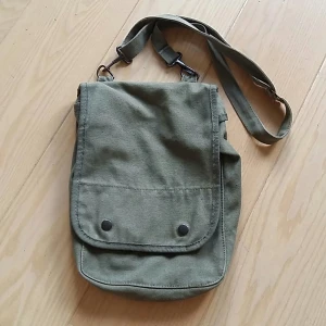 Väska i militär stil i kraftig bomull - En mindre väska i militär stil, militär grön, kraftig bomull. Metallic appar, tre avdelningar inne samt en liten ficka. Storlek ca 20*30 cm. Gott skick.