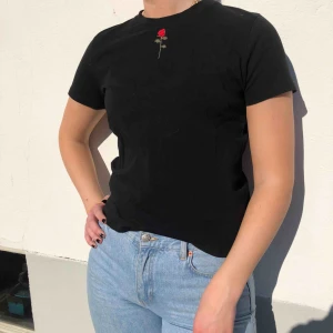  - Svart T-Shirt från NAKD med en ros som detalj på bröstet. Tröjan har bra kvalité och är enkel, snygg och passar till allt! 