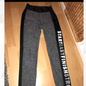  - Gråa träningsbyxor med svarta och vita detaljer. Text på vänstra benet. Resår i midjan