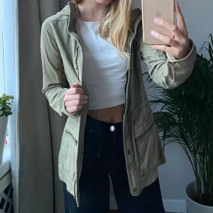 Hollister jacka strl XS - Snygg jacka från Hollister! Använd men fortfarande i mycket bra skick. Färgen är militärgrön, och tonen är en aning varmare än på bilderna. Plagget har både dragkedja och knappar, en ficka på vardera sida, samt ett hål i ena fickan för hörlurar (se bild 3). Köparen står för frakt! Startbud: 100kr 😽 >fråga gärna för fler bilder<     