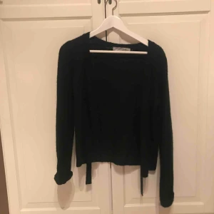  - Svart stickad cardigan i storlek M. Band som går att knyta till rosett uppe vid halsen. I fint skick 