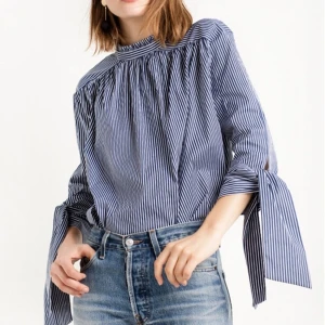 Randig skjorta - Skjorta i vit och blåa ränder med fina detaljer. Knäppning i rygg och knytning vid armslut. Aldrig använd, därför har jag produktbilder men jag kan även skicka bild på den på galge. 