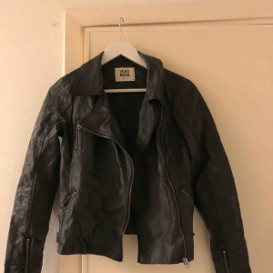 - Svart skinnjacka (ej äkta) med insytt rutigt mönster över ärmarna och silverdetaljer. Köparen står för frakten.