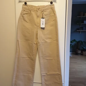 Beige byxor från Stradivarius - Helt nya beigea 90-talsjeans från Stradivarius. Finns i storlek 38 &36. Alla lappar sitter kvar. Rak modell i benen. Ej fållade nertill, avklippt kant. Snygg passform som är tight över midja/rumpa och lös över benen. HELT NYA. . Passar perfe