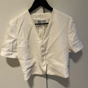 Vit kortärmad topp från Zara - Säljer en stilren vit kortärmad topp från Zara. Den har en v-ringad design med knappar framtill och är perfekt för både vardag och fest. Storlek M men passar även S