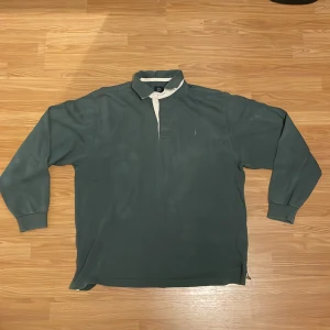 Grön pikétröja från Boomerang - Säljer en klassisk grön rugbytröja från Boomerang i storlek Large. Tröjan har långa ärmar och en vit krage som ger en snygg kontrast. Den är perfekt för både vardag och lite mer avslappnade tillfällen. 