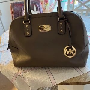 Brun handväska från Michael Kors - Säljer en snygg brun handväska från Michael Kors. Väskan är i skinn och har en stilren design med ett stort MK-hänge i guld. Den har dubbla handtag och en dragkedja upptill. Insidan är fodrad med Michael Kors-logotyp och har flera fack för enkel organisering. Perfekt för både vardag och fest!