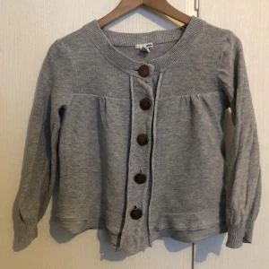 Grå kofta från H&M - Säljer en stilren grå kofta från H&M i storlek XS. Koftan har långa ärmar och knäpps med stora bruna knappar framtill. Den är tillverkad i en blandning av ramie och bomull, vilket ger en mjuk och bekväm känsla.