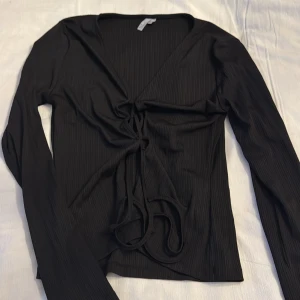 Svart ribbad topp med knytning - Snygg svart ribbad topp med långärmad design och knytning framtill. Perfekt för en stilren look.