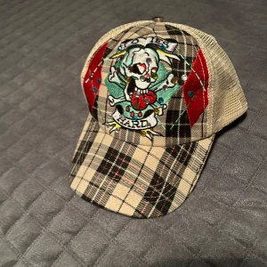 Rutig keps med broderad dödskalle - Snygg rutig keps med ett coolt broderi av en dödskalle och texten 'Ed Hardy' framtill. Kepsen har en klassisk skärm och justerbar passform. Perfekt för att ge din outfit en edgy touch.
