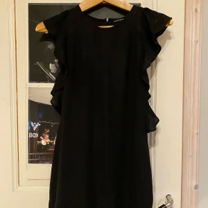 Svart klänning med volangdetaljer - Elegant svart klänning med volangdetaljer längs sidorna. Perfekt för en stilren look. Klänningen har en rund hals och är ärmlös, vilket ger en modern och chic känsla.