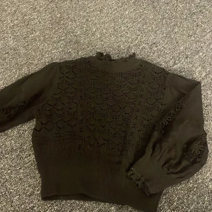 Svart stickad tröja med spetsdetaljer - Elegant svart stickad tröja med vackra spetsdetaljer på fram- och baksidan. Tröjan har långa ärmar med ribbade muddar och en hög halsringning. Perfekt för kyligare dagar. Jätte fin med ett vitt linne under 