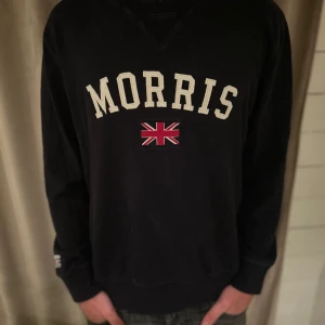 Svart sweatshirt från Morris - Tjena, säljer enSnygg mörkblå sweatshirt från Morris med vit text och en Union Jack-detalj på framsidan. Tröjan har långa ärmar och en klassisk passform. Perfekt för en avslappnad stil.