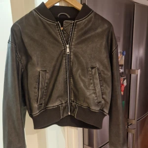 Svart bomberjacka i skinnimitation - Snygg svart bomberjacka i skinnimitation med dragkedja framtill och ribbade muddar. Jackan har en klassisk bomberstil med två fickor och en bekväm passform. Perfekt för en cool och avslappnad look.