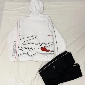 Vit lacoste tracksuit  - Snygg vit vindjacka från Lacoste med dragkedja och huva. Jackan har ett stort krokodilmotiv och texten 'Lacoste' längs ena ärmen. Perfekt för en sportig look. Storlek S-M skicket 10/10 helt ny 