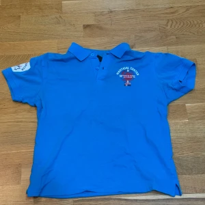 Blå pikétröja från Cross Sportswear - Snygg blå pikétröja från Cross Sportswear med 'Nautical Center' tryck på bröstet och ärmen. Perfekt för en sportig look med maritimt tema. Tröjan har korta ärmar och en klassisk krage med knappar.