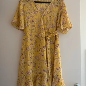 Gul blommig klänning från Amelie & Me - Charmig gul klänning med blommigt mönster från Amelie & Me. Klänningen har korta ärmar och en omlott-design med knytband i midjan. Perfekt för en somrig look!