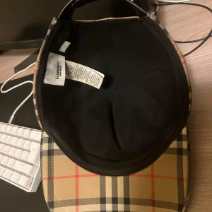 Rutig keps från Burberry - Snygg rutig keps från Burberry med klassiskt mönster i beige, svart och rött. Kepsen har justerbar rem baktill för perfekt passform. Perfekt accessoar för en stilren look. Tar bara emot byten kan mötas i skövde eller Hjo 