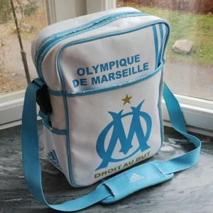 Vit och blå axelväska från Olympique de Marseille - Säljer en vit och blå axelväska med Olympique de Marseille-logga. Väskan har en justerbar axelrem och dragkedja upptill. Perfekt för fotbollsfans som vill visa sitt stöd för laget.