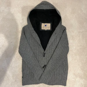 Fodrad Cardigan - Säljer en snygg grå ribbad kofta med huva och knappar framtill. Perfekt för kyliga dagar med sitt mjuka foder. Material: Ull. Koftan har långa ärmar och en stilren design som passar till det mesta. Vid minsta fråga eller fundering hör av er🤝