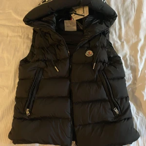 Svart dunväst från Moncler - Snygg svart dunväst från Moncler med dragkedja och broderad logotyp på bröstet. Västen ett vitt moncler tryck på luvan som även går att gömma med hjäp av dragkedja. Hör av dig vid intresse!! 