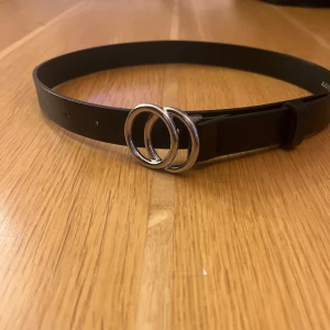 Svart bälte från Lindex - Snyggt svart bälte från Lindex med dubbla silverringar som spänne. Perfekt för att ge en stilren touch till din outfit. och mäter 65 cm i längd.
