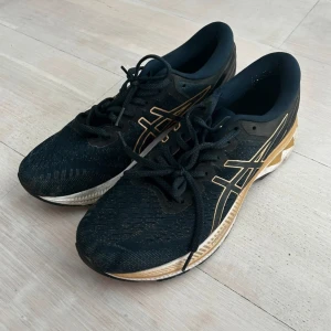 Svarta och guldiga Asics löparskor - Säljer ett par svarta Asics löparskor med guldiga detaljer. Skorna har snörning och en slitstark sula, perfekt för löpning och träning.