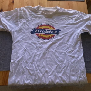 Vit t-shirt från Dickies - Säljer en klassisk vit t-shirt från Dickies med deras ikoniska logga i rött, gult och blått på bröstet. Perfekt för en avslappnad stil.