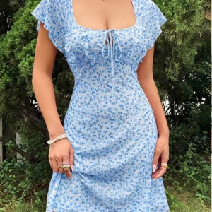 Blå blommig klänning - Fin blå klänning med blommigt mönster och korta ärmar. Den har en söt knytning framtill och en figurnära passform som framhäver silhuetten🖤