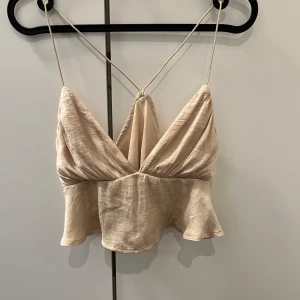 Beige linne med tunna axelband - Snyggt beige linne med tunna axelband och djup v-ringning. Toppen har en luftig passform och korsade band i ryggen, vilket ger en stilren look. Perfekt för varma sommardagar.