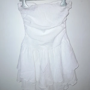 Vit axelbandslös klänning - Söt vit axelbandslös klänning med volanger. Perfekt för somriga dagar eller festliga tillfällen. Den har en smickrande passform och är tillverkad i ett lätt och luftigt material.